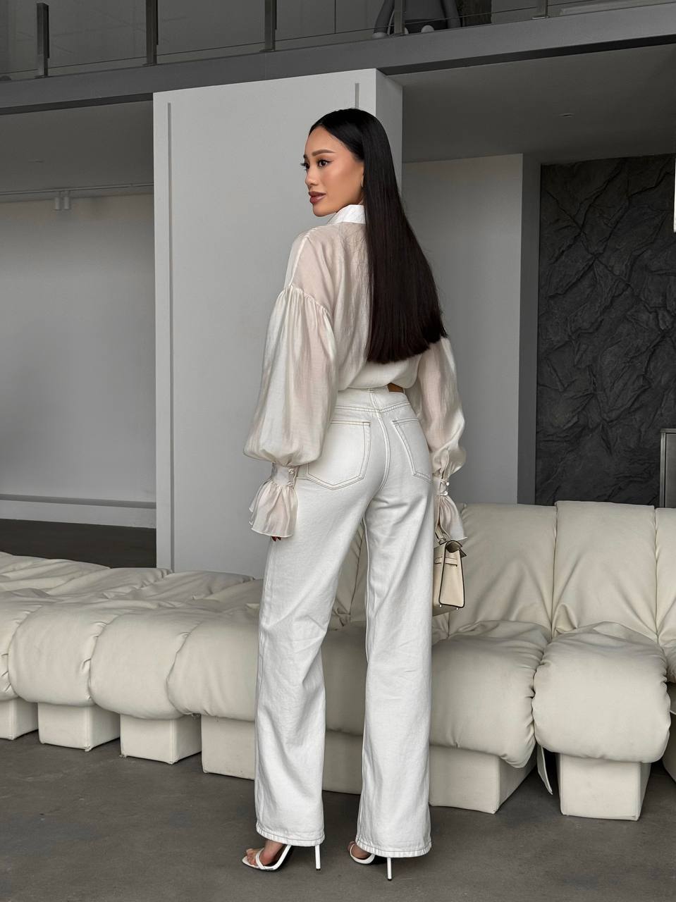 Woman in a white outfit standing in a modern room with gray walls and a white sofa.