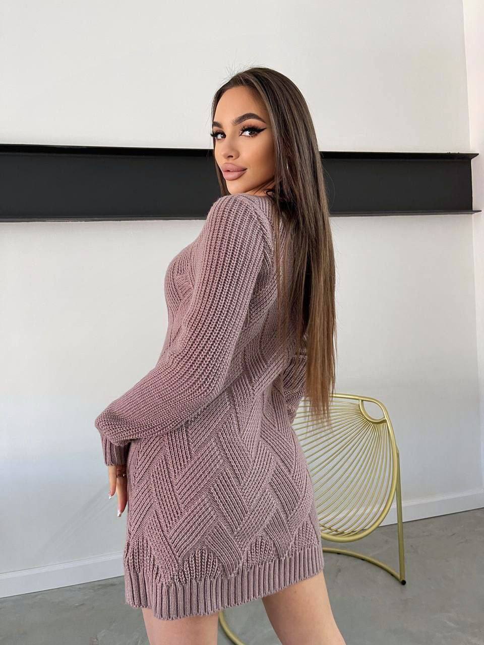 Woman wearing a pink knit dress standing in a minimalistic room with a white wall and black shelf.