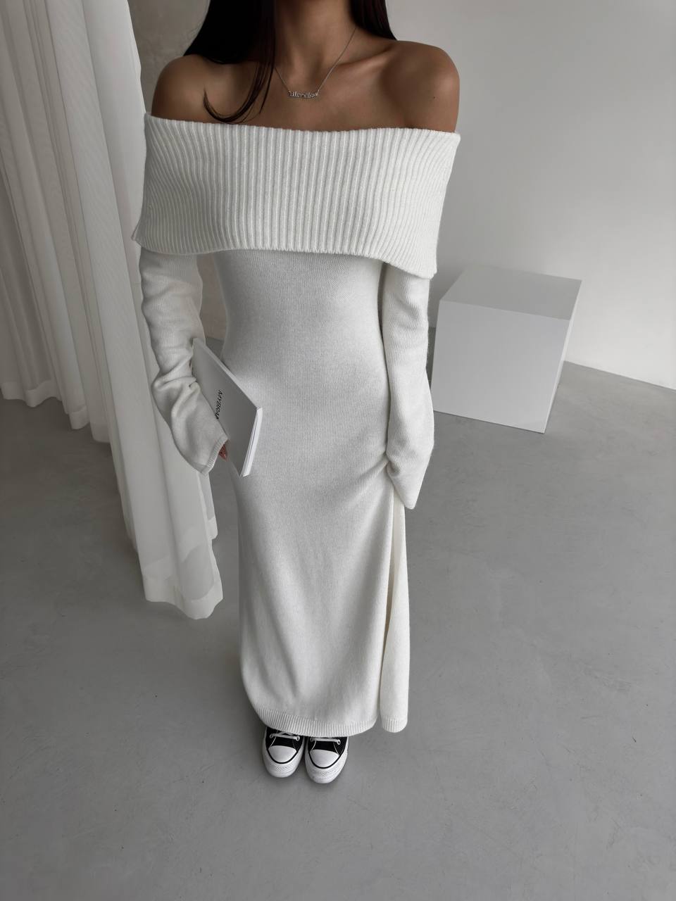 Person wearing a white off-shoulder top and light gray pants in a minimalistic setting.