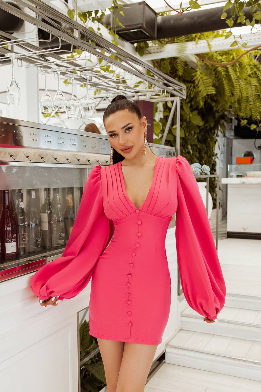 Woman in a bright pink dress standing in an outdoor setting with greenery and a building in the background.