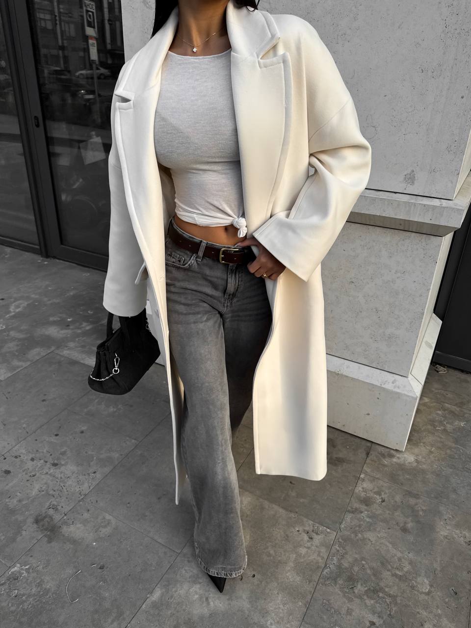 Person wearing a long cream coat over a gray top and jeans, standing on a sidewalk.