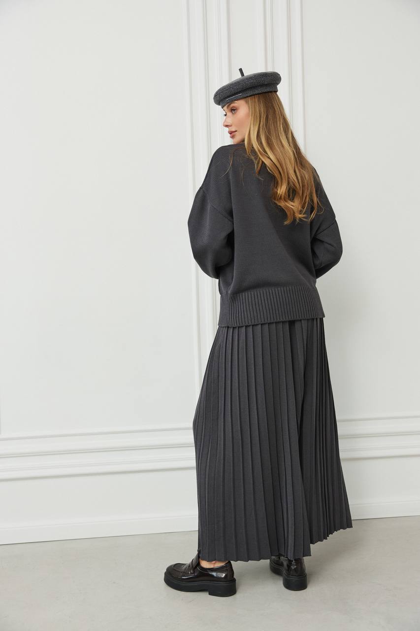 Person wearing a dark gray outfit with a pleated skirt and beret against a white wall.