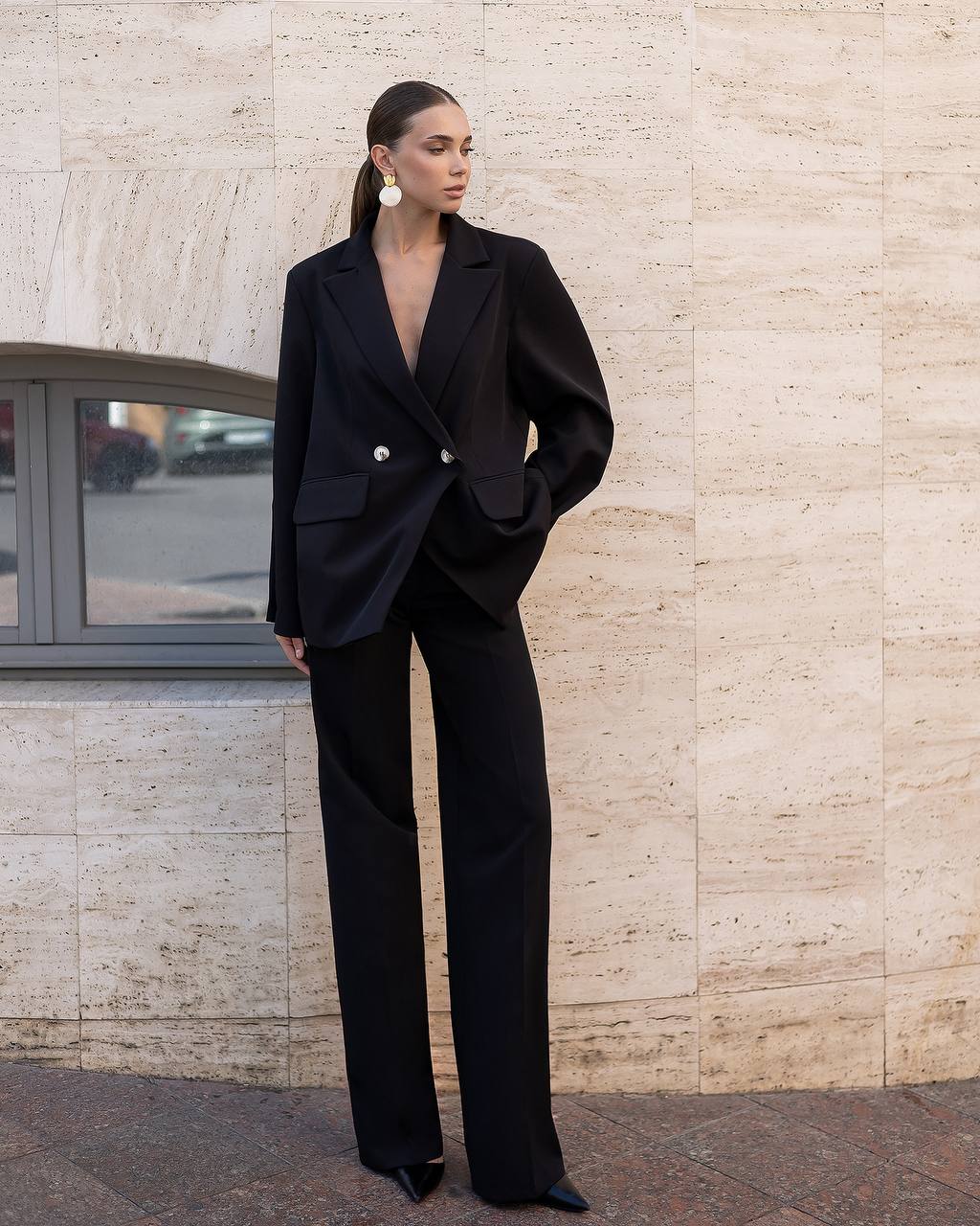 A woman stands outdoors against a beige stone wall wearing an oversized black double-breasted blazer with wide-leg black trousers, black pointed shoes, and large white hoop earrings.