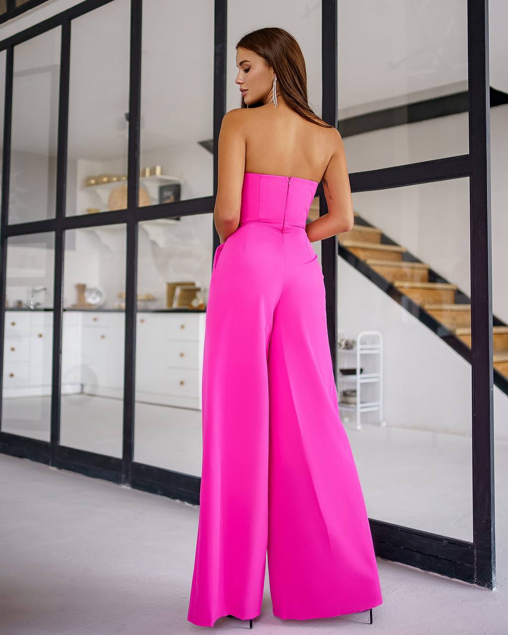 Woman wearing a bright pink strapless jumpsuit in a modern interior setting.