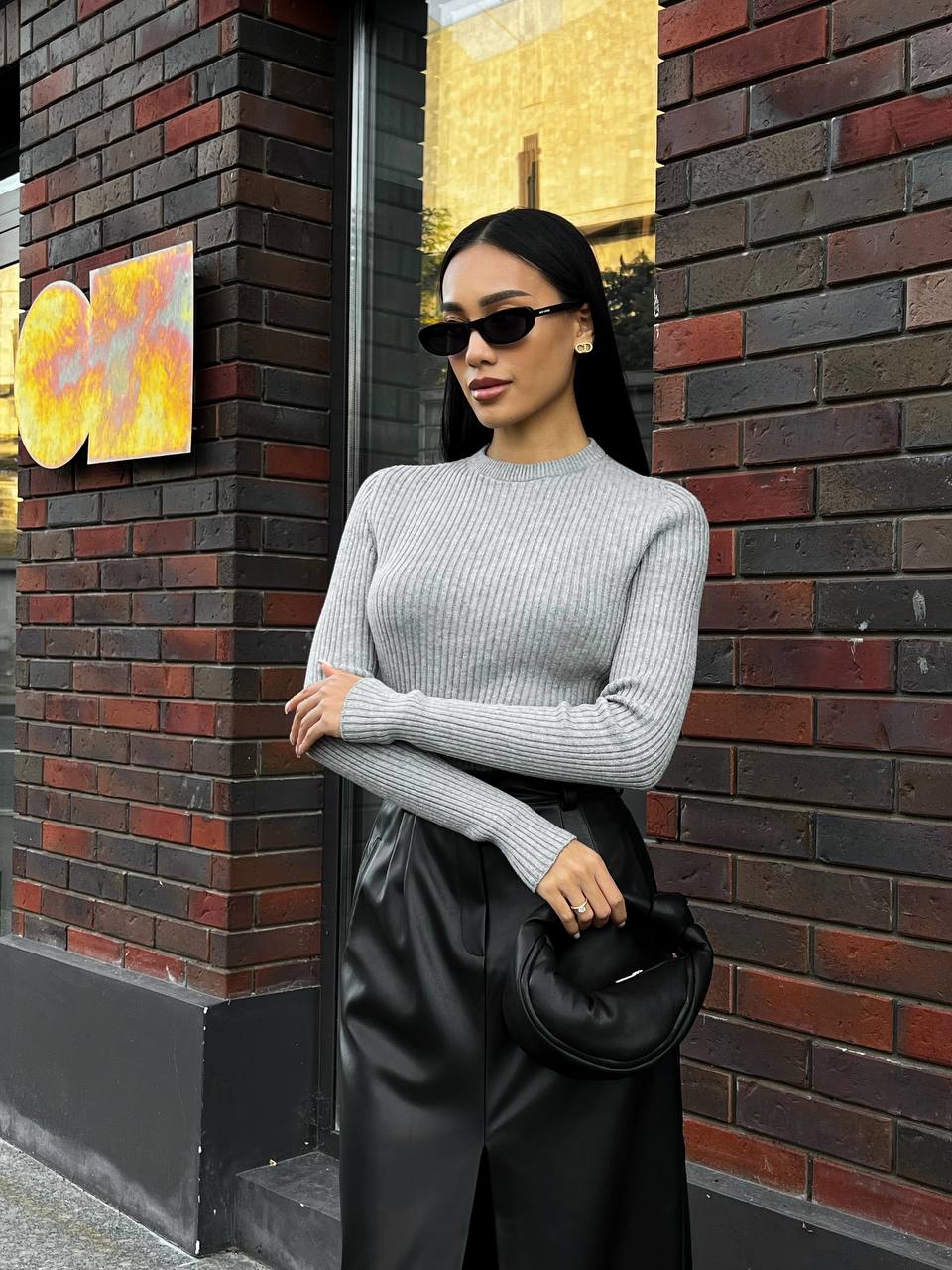 Woman in a gray sweater and black pants standing against a brick wall.