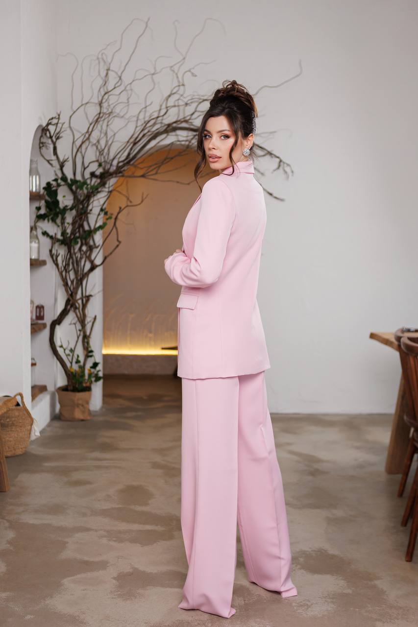 Woman in a pink outfit standing in a room with a plant and wooden furniture.