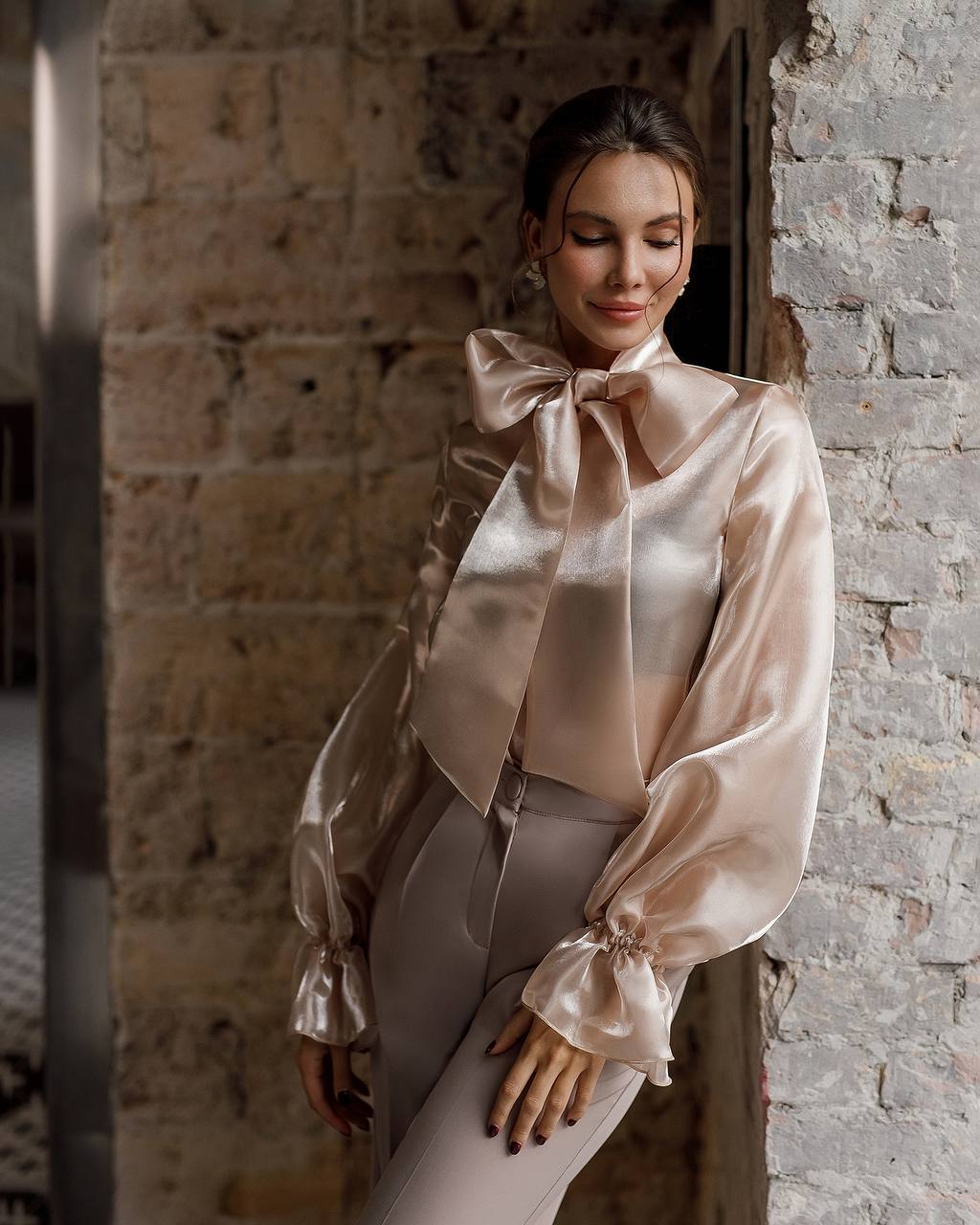 Woman wearing a beige satin blouse with a large bow tie against a stone wall.