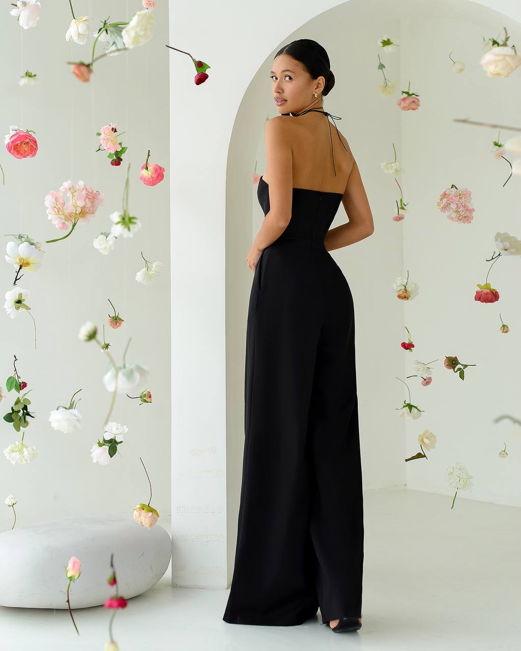 Woman in a black jumpsuit standing in a room with floral decorations on the walls.