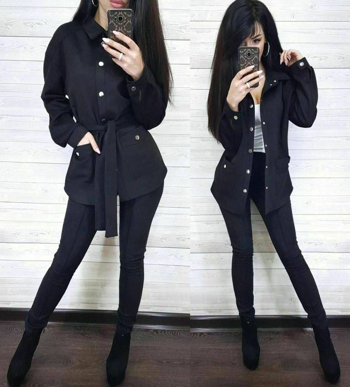 Person wearing a black outfit with a belt, standing in front of a white wall.