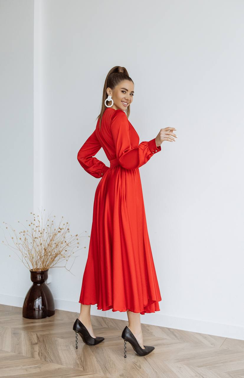 Woman in a red dress standing against a white wall with a vase of dried plants on a wooden floor.