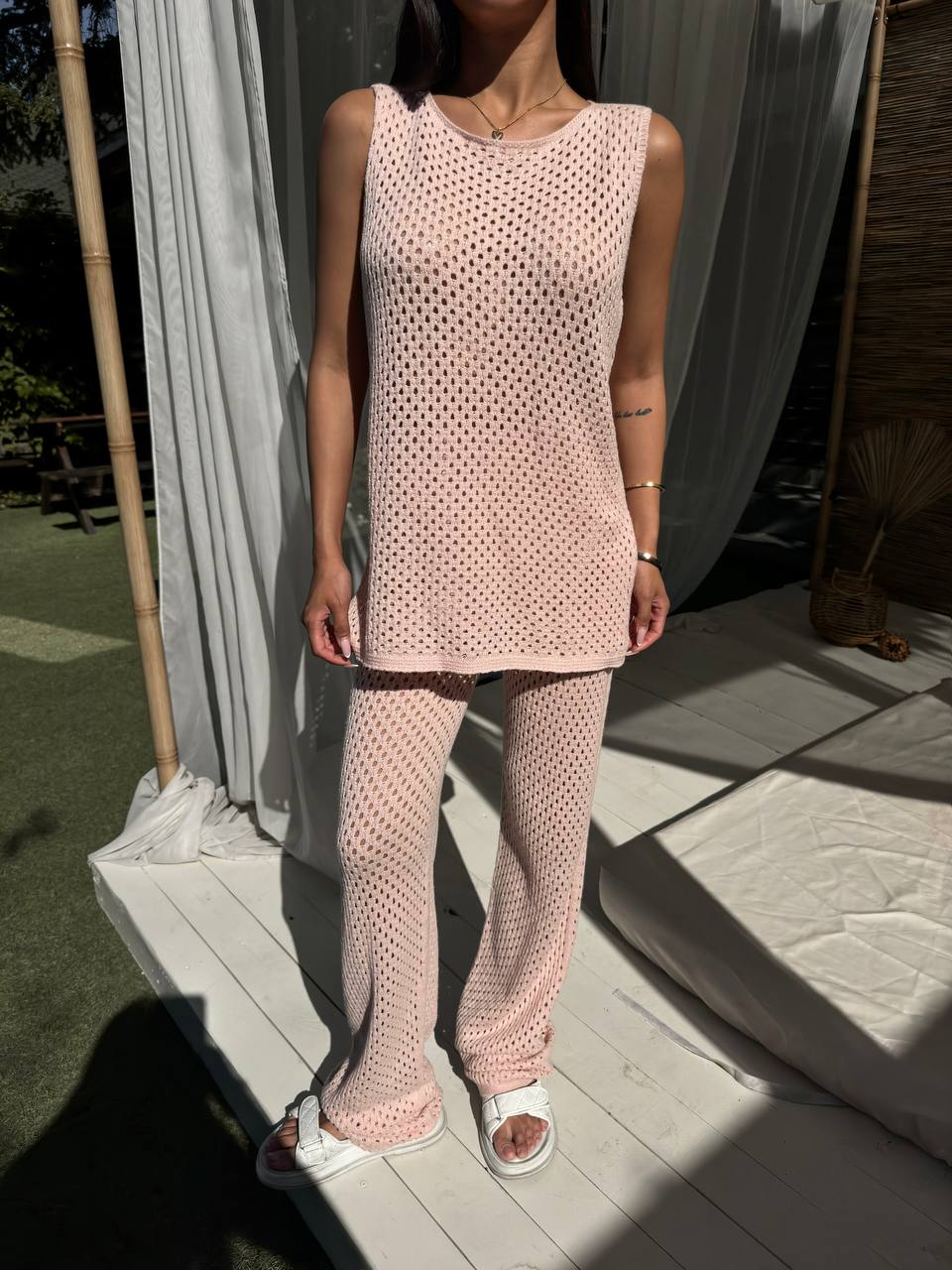 Person wearing a pink knitted outfit standing outdoors with light-colored curtains in the background.