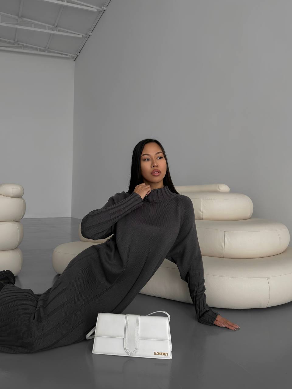 Woman in a dark outfit sitting on a white handbag with a minimalistic background