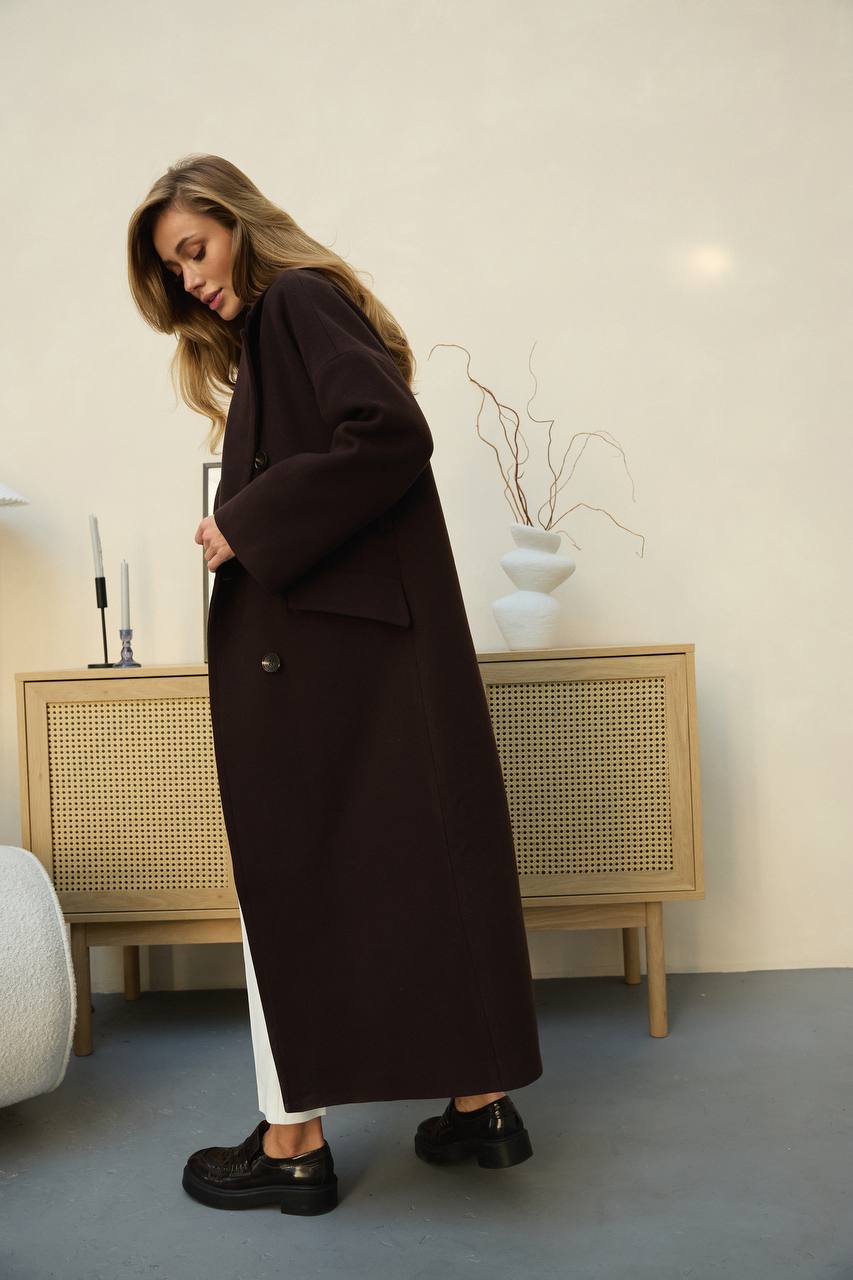 Woman wearing a long dark coat in a minimalistic room with a wooden cabinet and white walls.