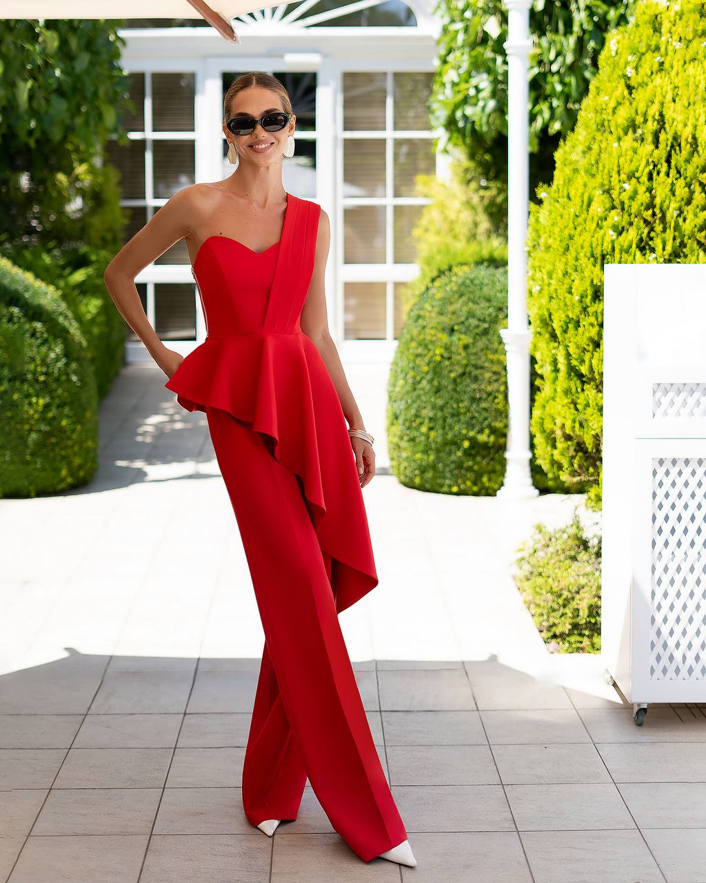 Woman in a red one-shoulder jumpsuit standing outdoors with greenery in the background