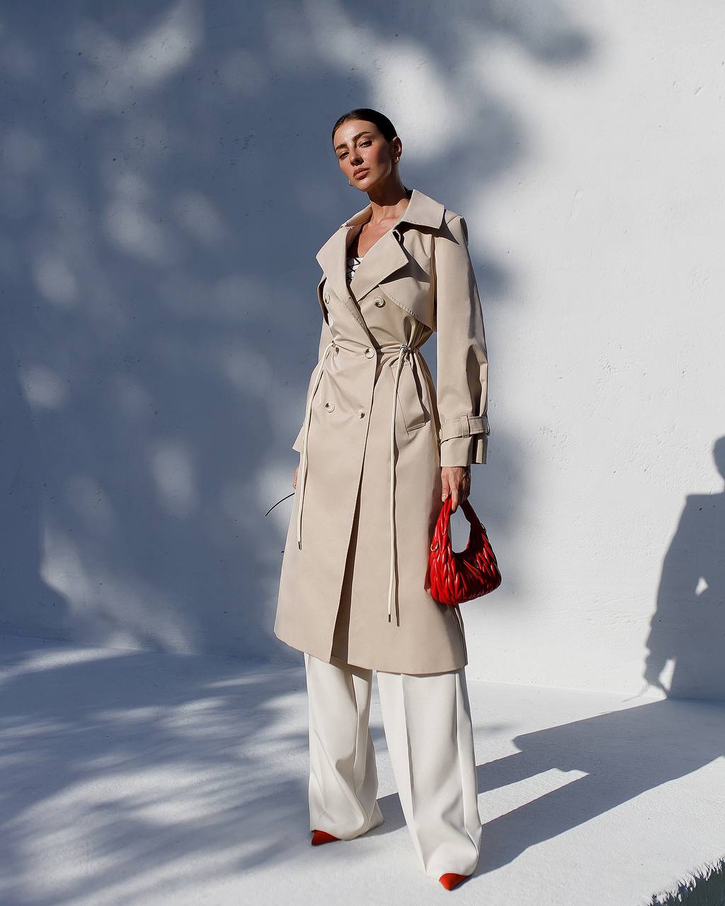 Woman wearing a beige trench coat and white pants, holding a red handbag against a light background.