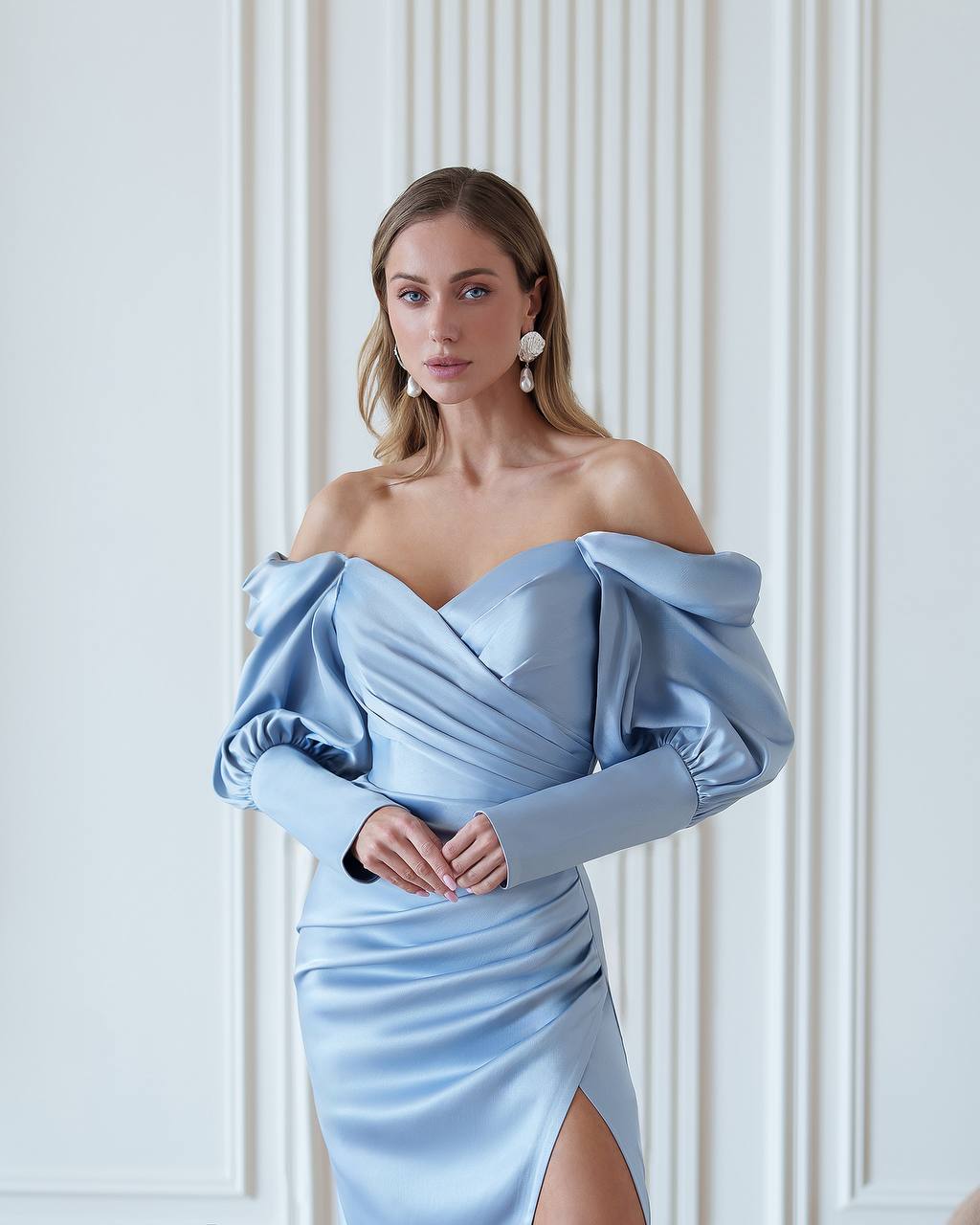 Woman wearing a light blue off-shoulder dress against a white wall.