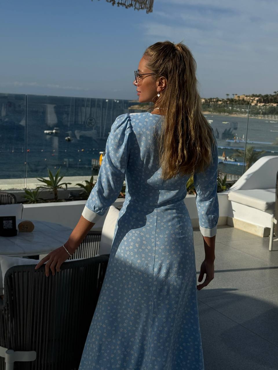Woman in a blue dress standing on a balcony with a scenic view of water and buildings.
