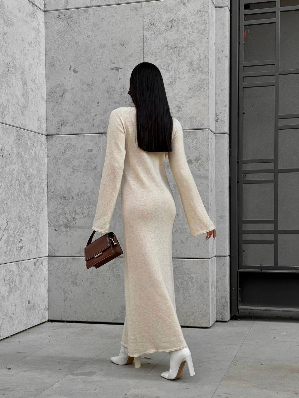 Woman in a long beige dress walking away from a gray building.