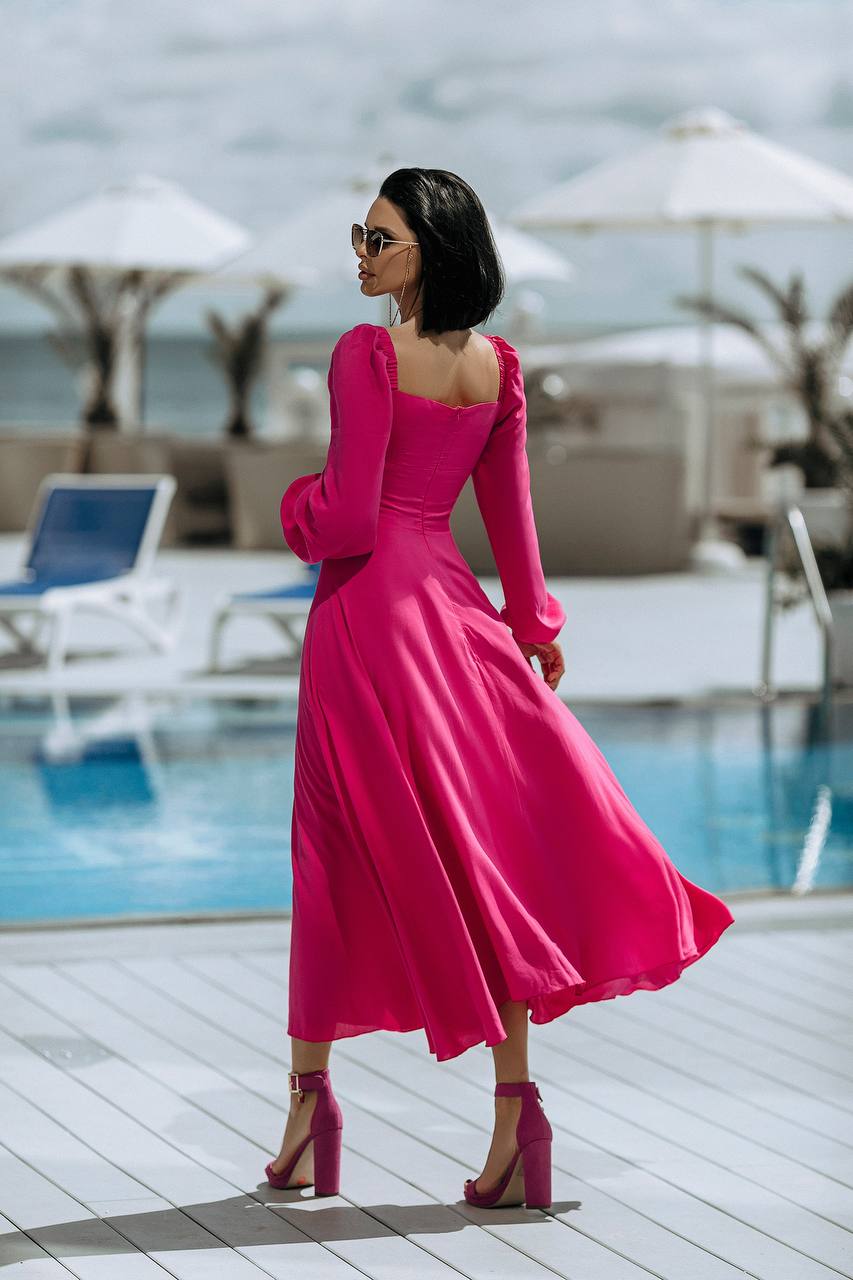 Woman in a pink dress standing by a poolside with umbrellas and lounge chairs in the background.