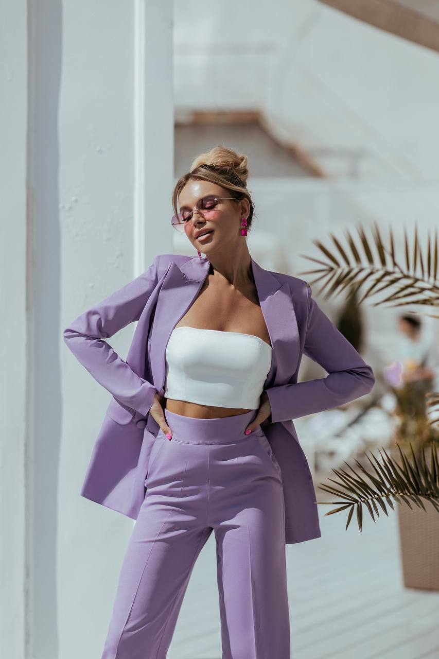 Woman in a purple suit and white top standing outdoors with plants in the background