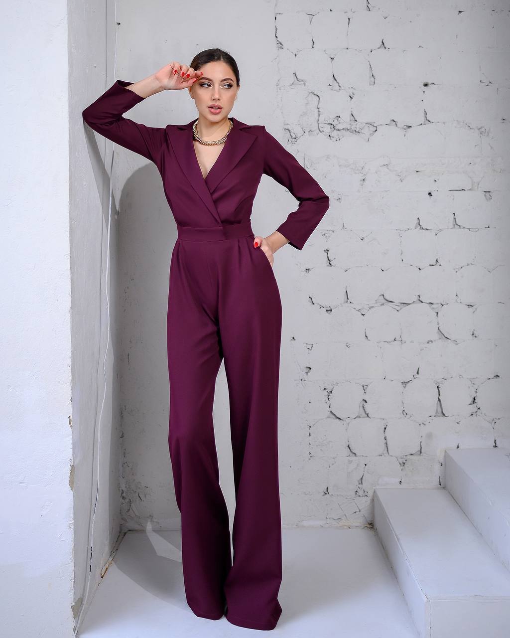 Woman wearing a burgundy jumpsuit against a white textured wall.