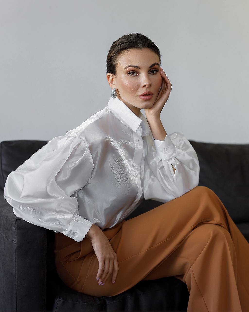 Woman wearing a white blouse and brown pants sitting on a black couch against a gray background