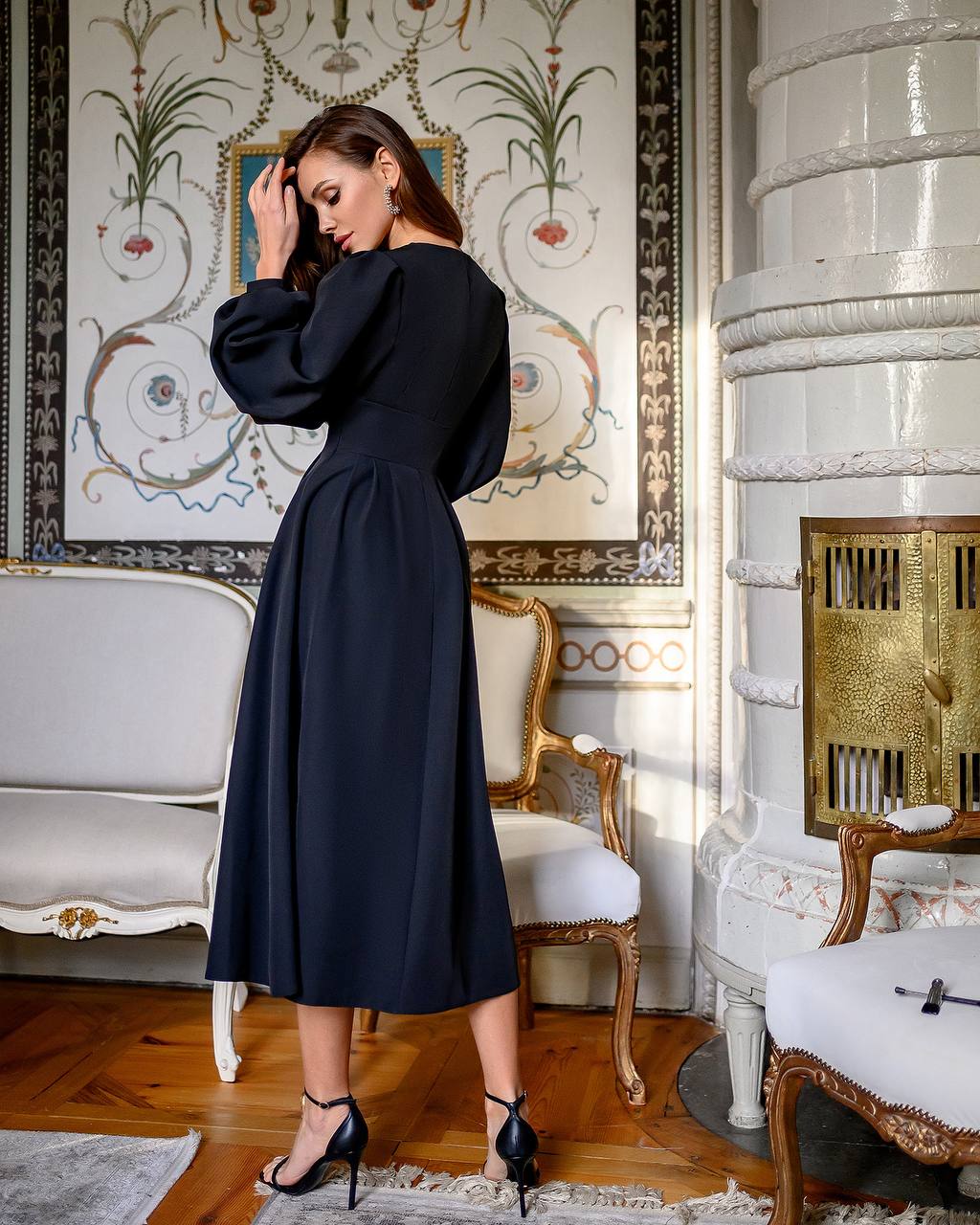Woman in a black dress standing in an ornate room with decorative wallpaper and furniture.