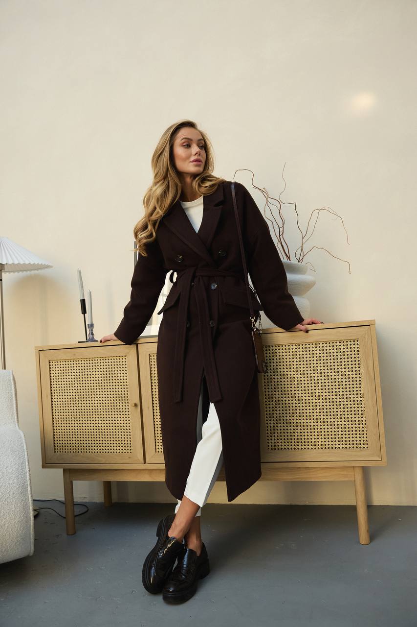 Woman in a long dark coat standing next to a wooden cabinet in a room.