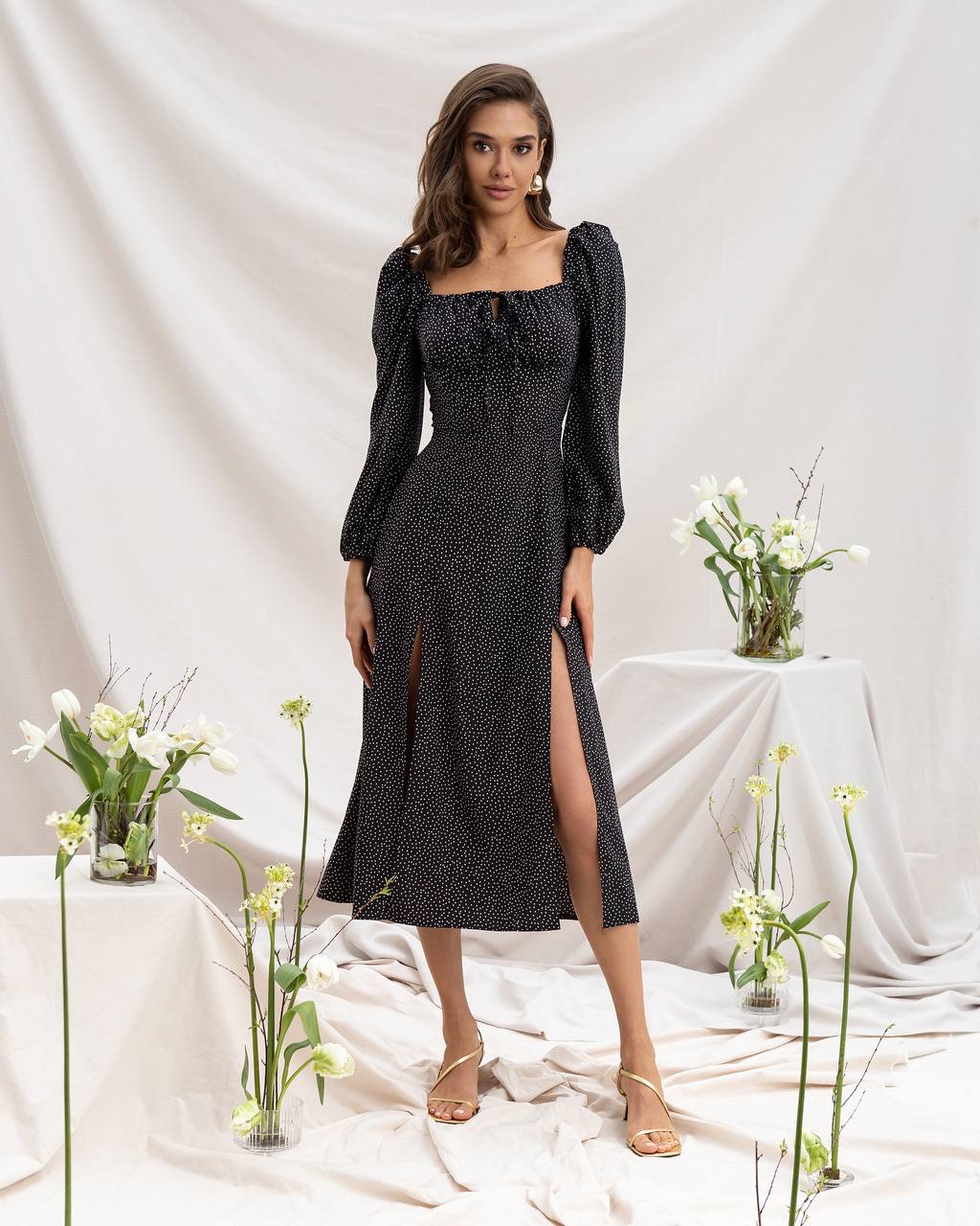 Woman wearing a black patterned dress with long sleeves and a slit, standing in a softly lit room with white draped fabric and floral arrangements.