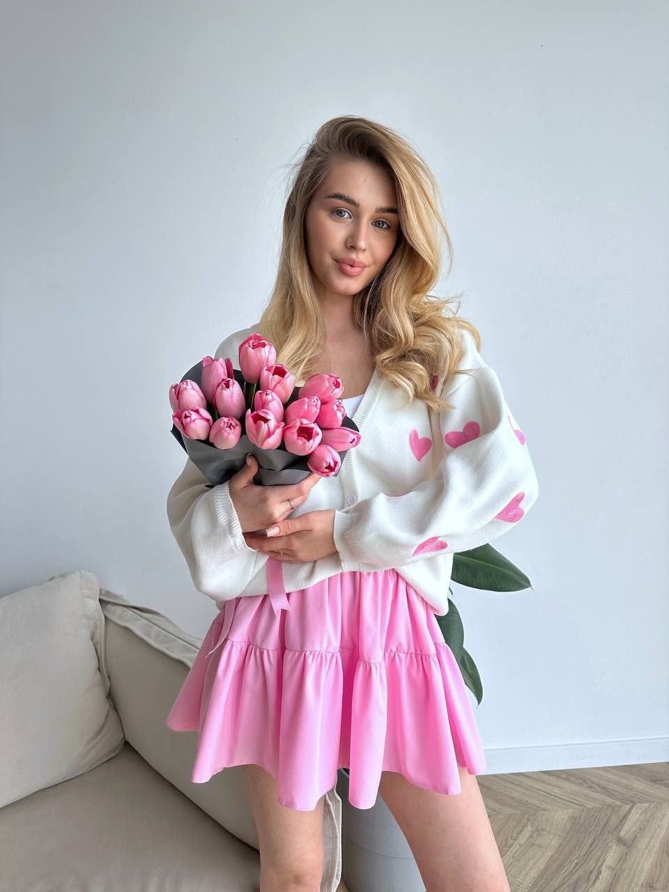Woman holding a bouquet of pink tulips wearing a white sweater with pink heart patterns and a pink skirt.
