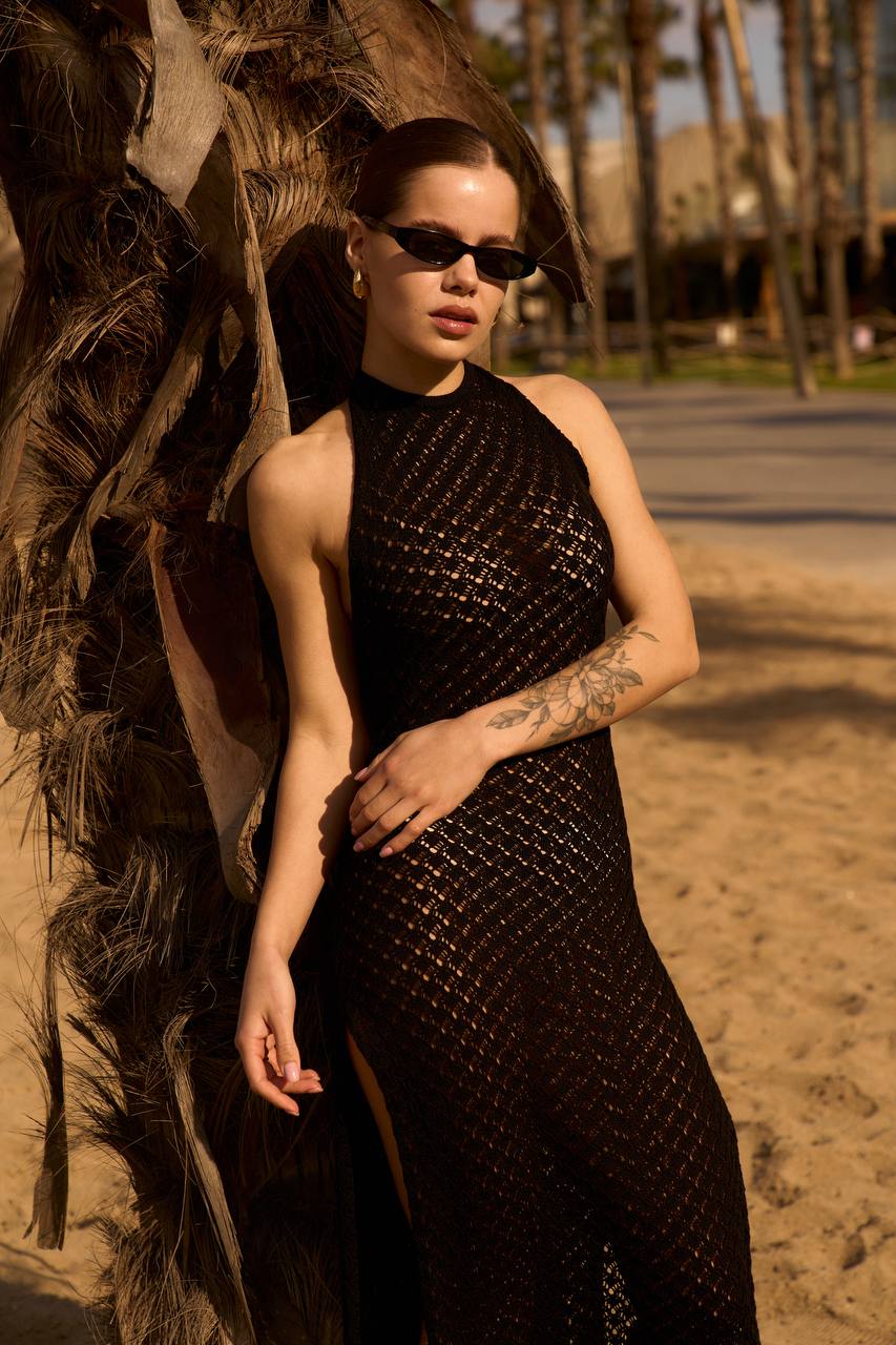 Woman in a black dress standing next to a palm tree on a beach.