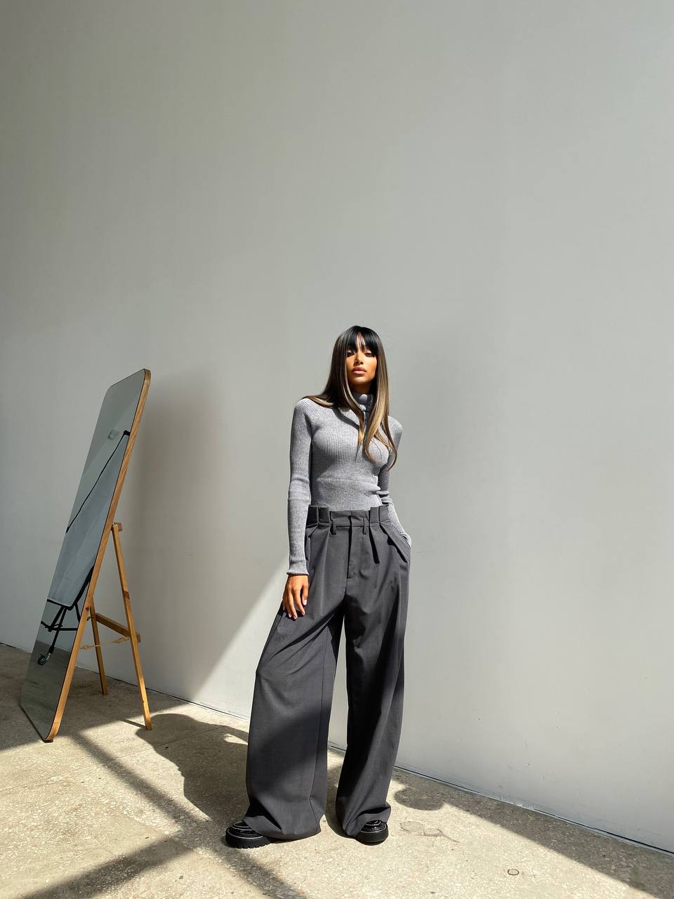 Person wearing a gray outfit standing against a plain wall with a mirror on an easel.