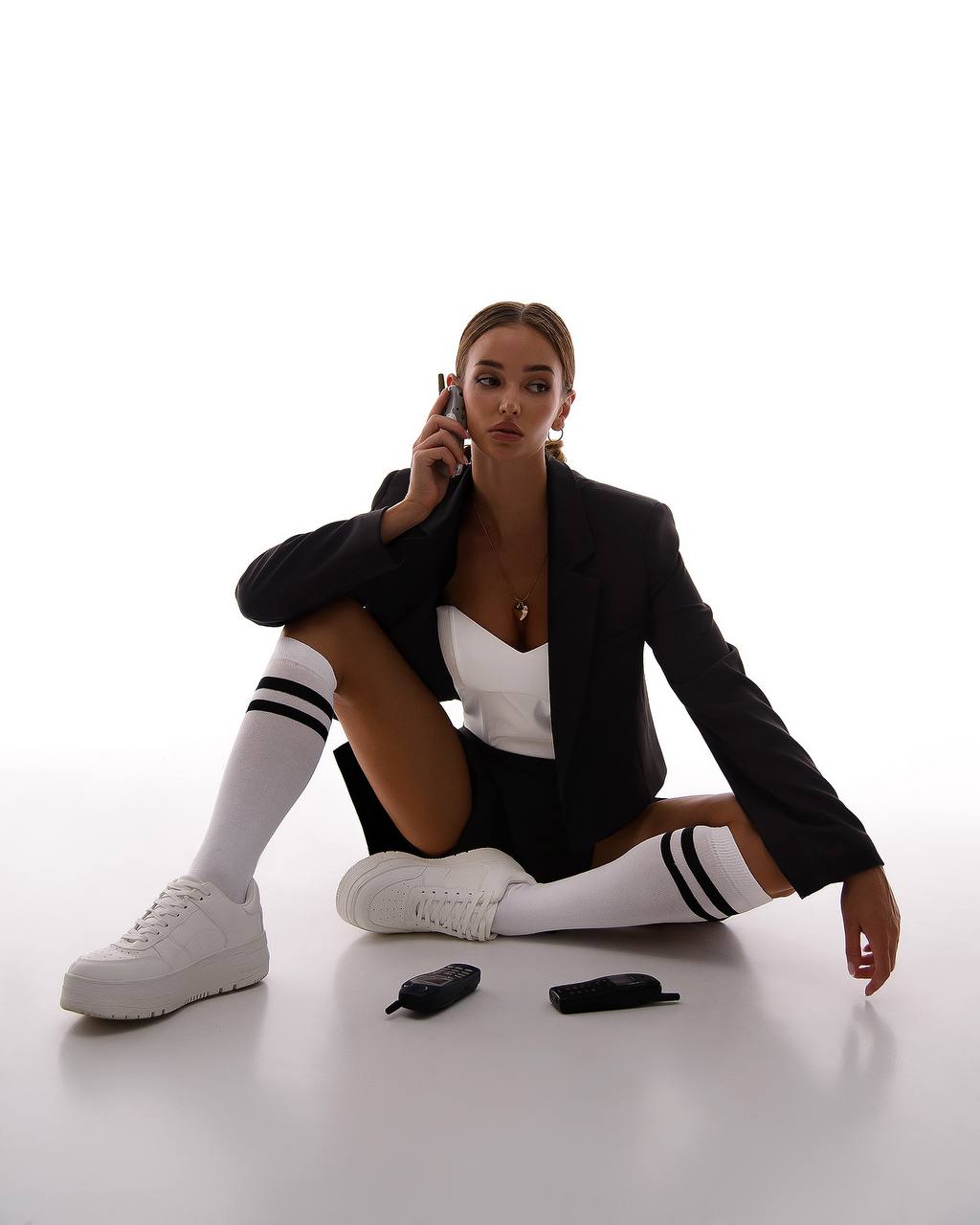 Woman in a black blazer and white outfit sitting on the floor with white sneakers and knee-high socks.