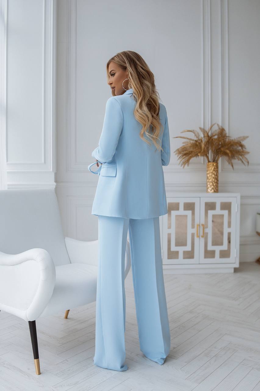 Woman in a light blue suit standing in a stylish room with white walls and decor.