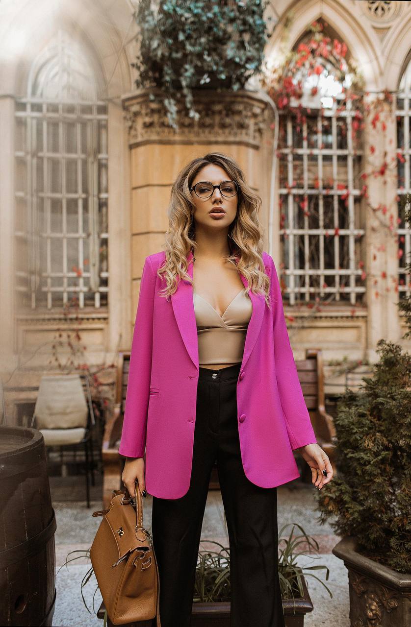 Woman wearing a bright pink blazer over a beige top in an outdoor setting with architectural elements.