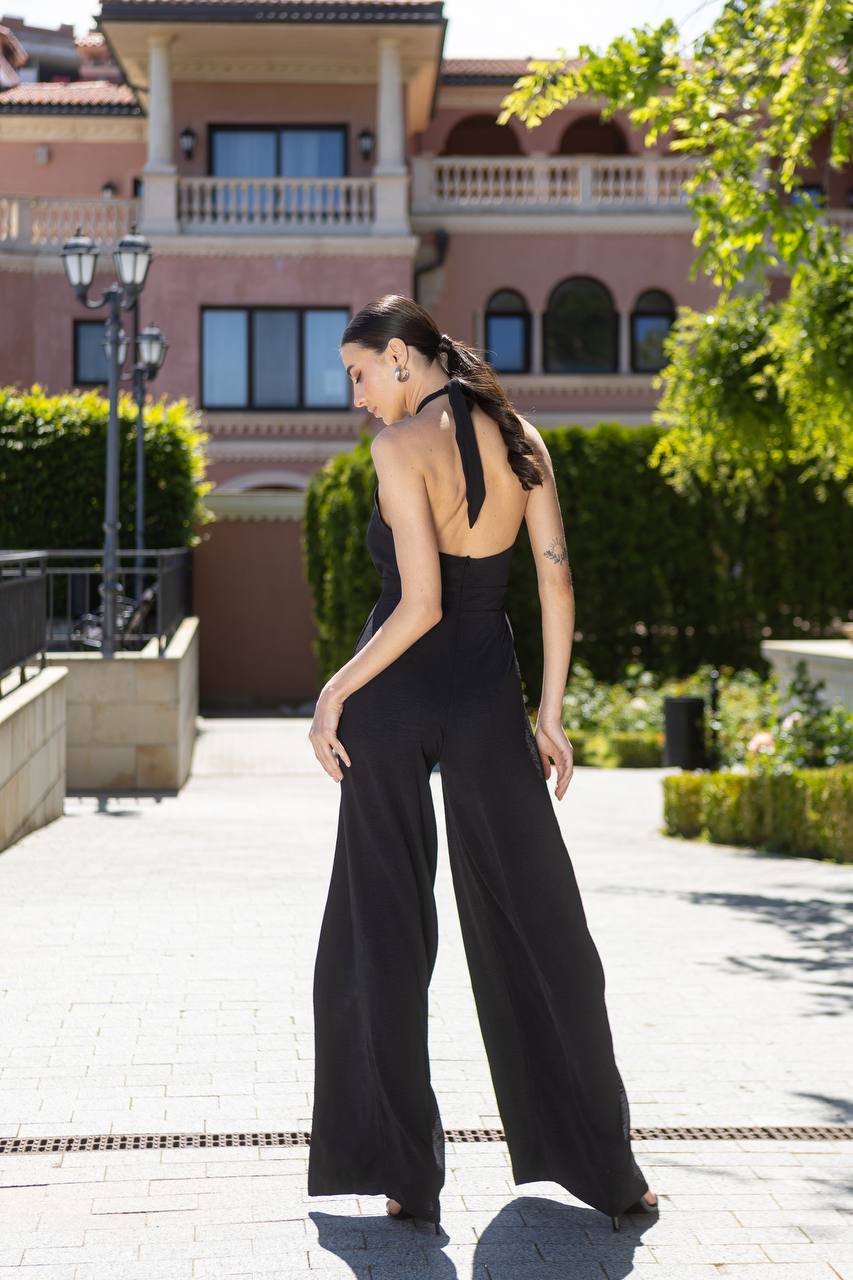 Woman in a black jumpsuit standing outdoors with a building and greenery in the background