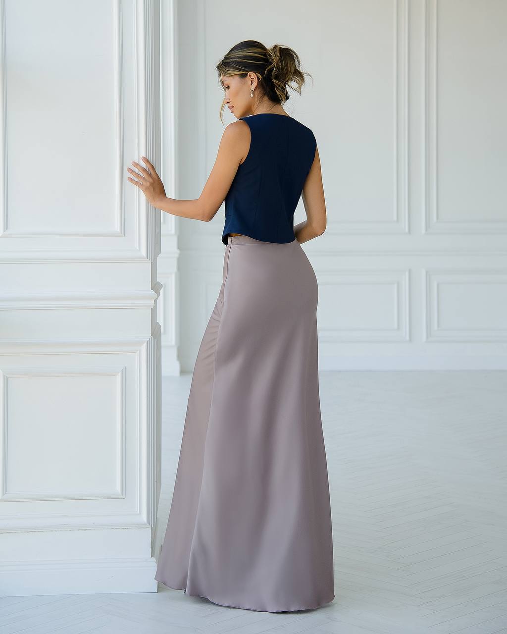Woman wearing a navy top and long light purple skirt against a white paneled wall.