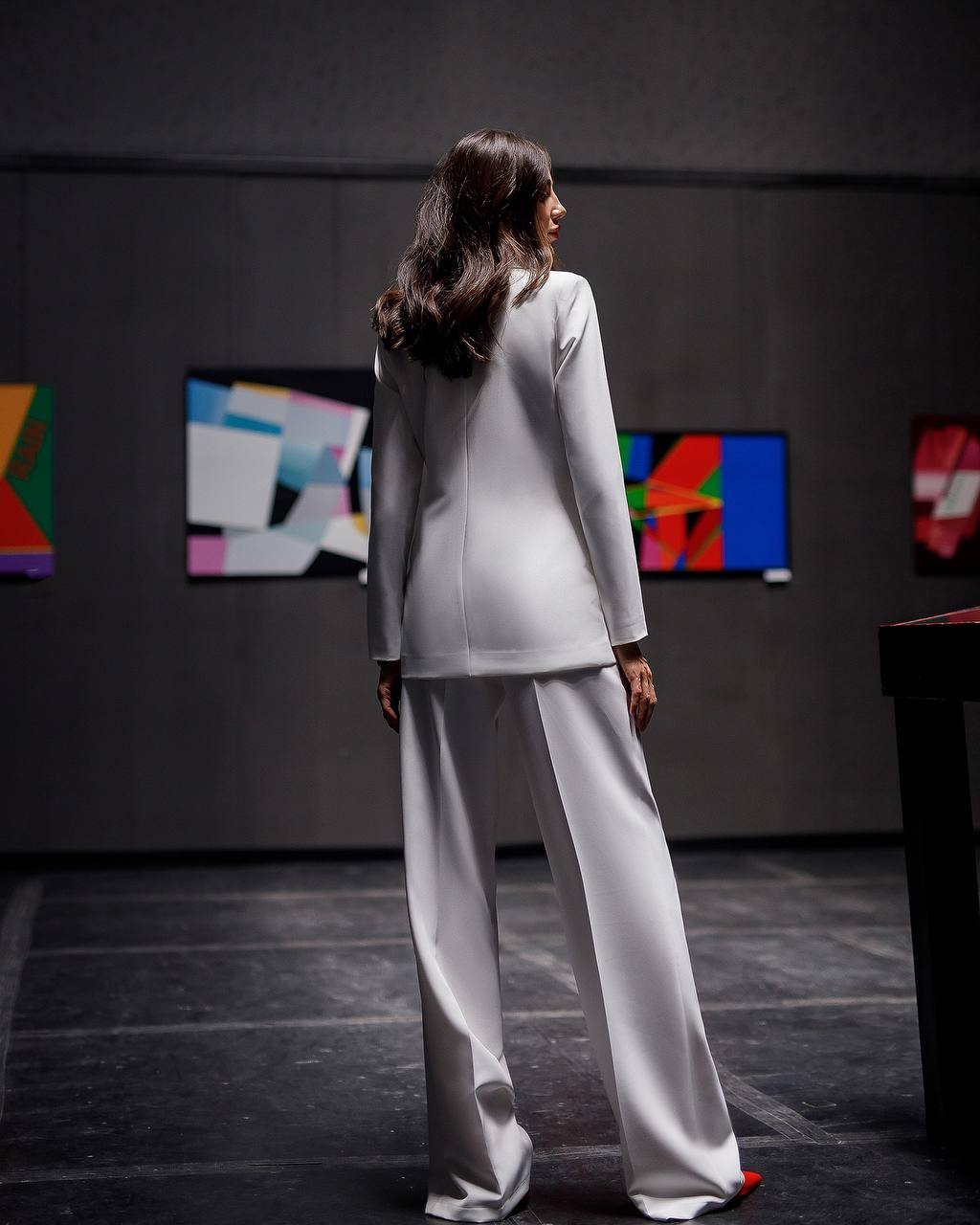 Woman in a white suit standing in front of colorful abstract paintings on a dark wall.