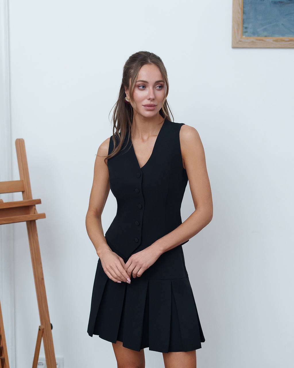 Woman wearing a black sleeveless dress standing against a white wall.
