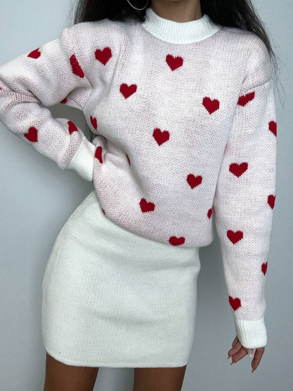 Person wearing a pink sweater with red heart patterns and a white skirt against a gray background