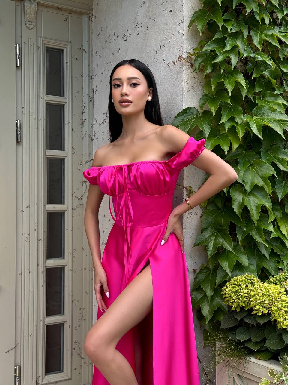Woman in a bright pink off-shoulder dress standing outdoors next to a building with greenery.