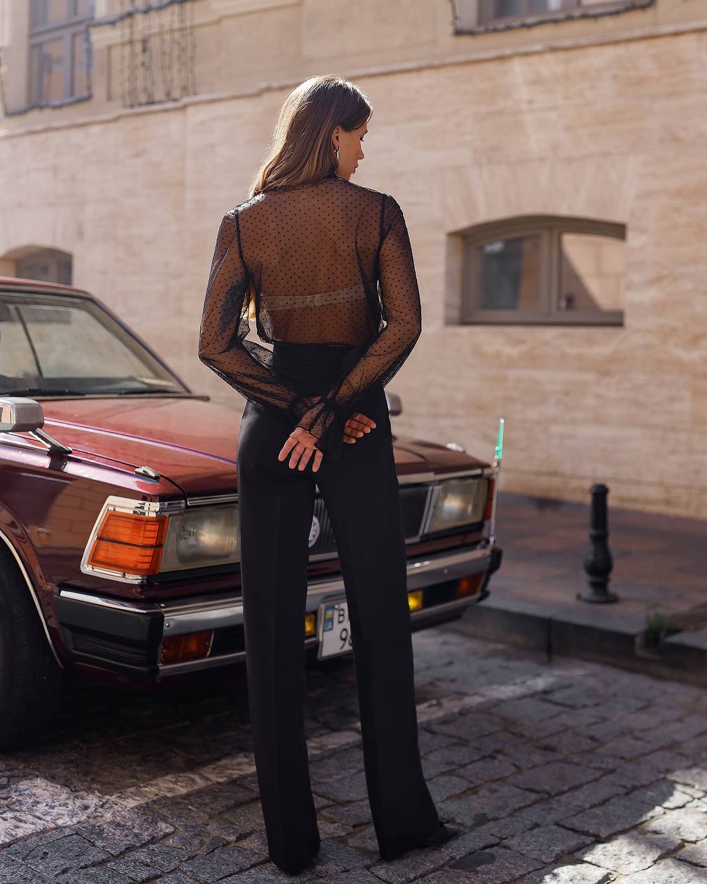 Woman in a black outfit standing next to a vintage car on a street.