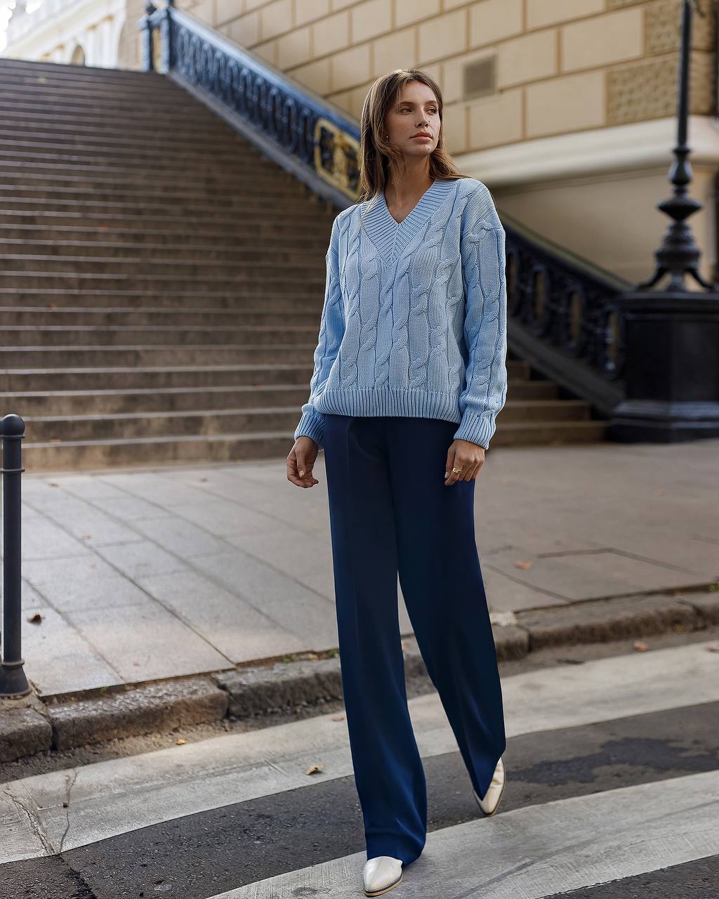 Woman wearing a blue cable knit sweater and dark blue pants walking on a city street.