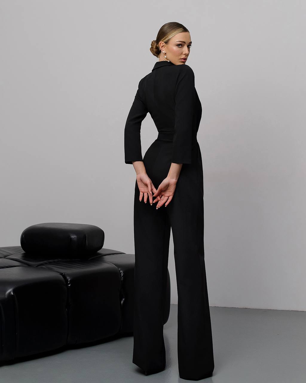 Woman wearing a black jumpsuit standing in a minimalistic room with a black couch.