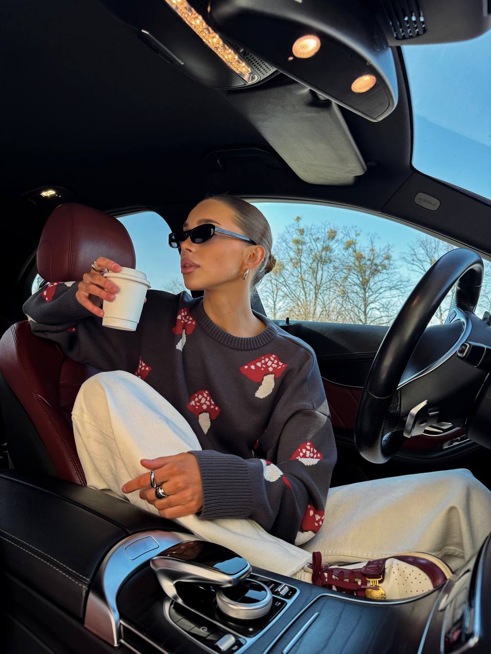 Woman sitting in a car holding a coffee cup, wearing a sweater with mushroom designs.