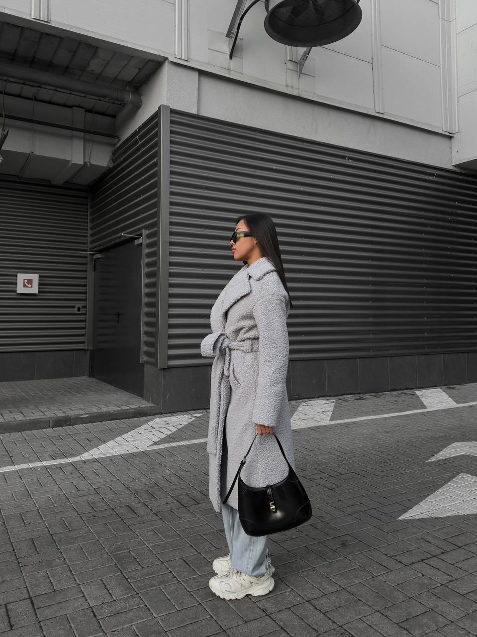 Person wearing a long gray coat and light blue jeans standing on a street.