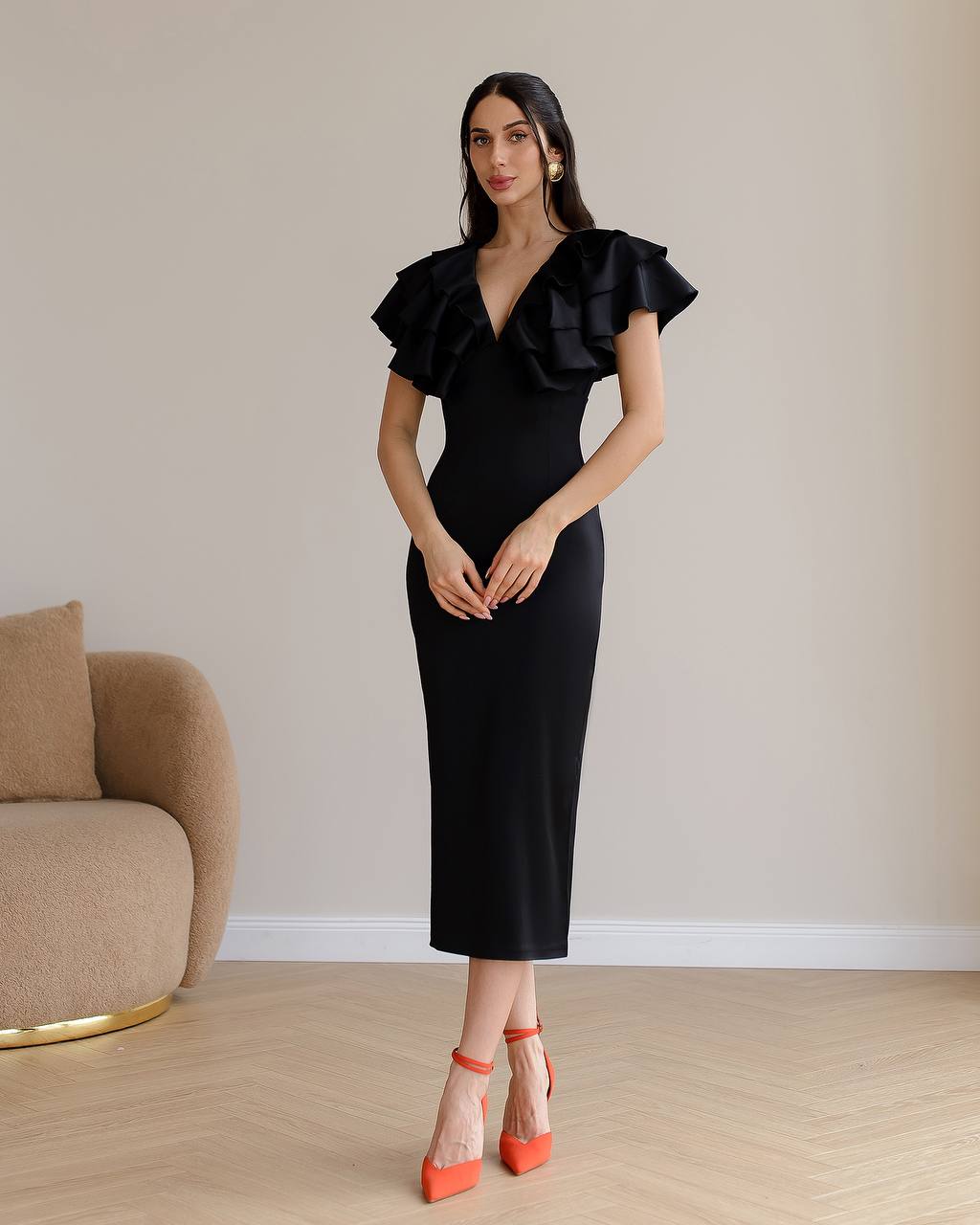 Woman wearing a black dress with ruffled sleeves in a room with a beige wall and brown chair.