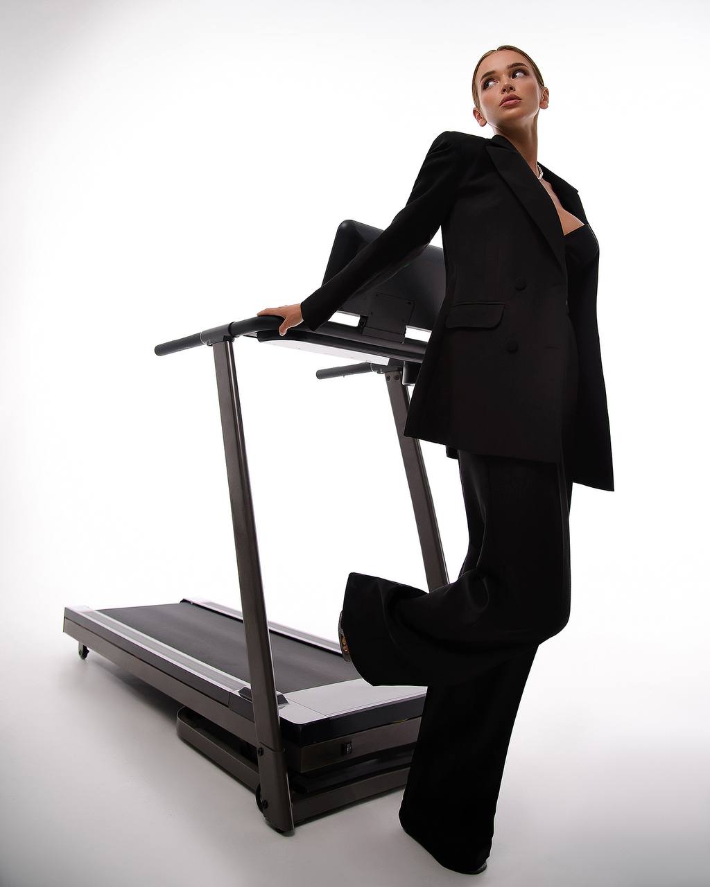 Woman in a black suit standing next to a treadmill on a white background