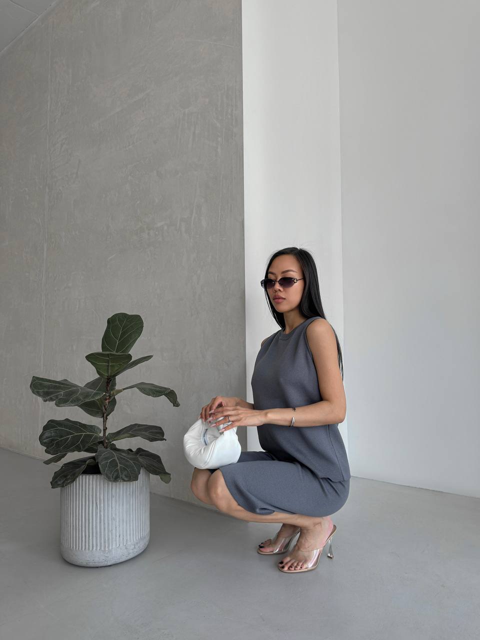 Woman in a gray dress holding a white vase next to a potted plant in a minimalistic setting.