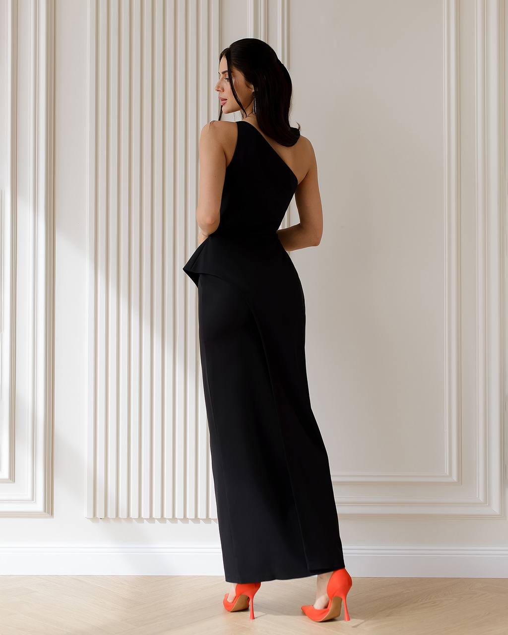 Woman wearing a black evening gown with a high slit, standing against a light-colored paneled wall.